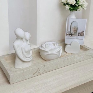 Op de afbeelding: Witte keramische schaal met een wit keramisch beeld van twee figuren die elkaar omhelzen, een witte keramische roos en een wit keramisch hart. De schaal staat op een licht houten oppervlak.