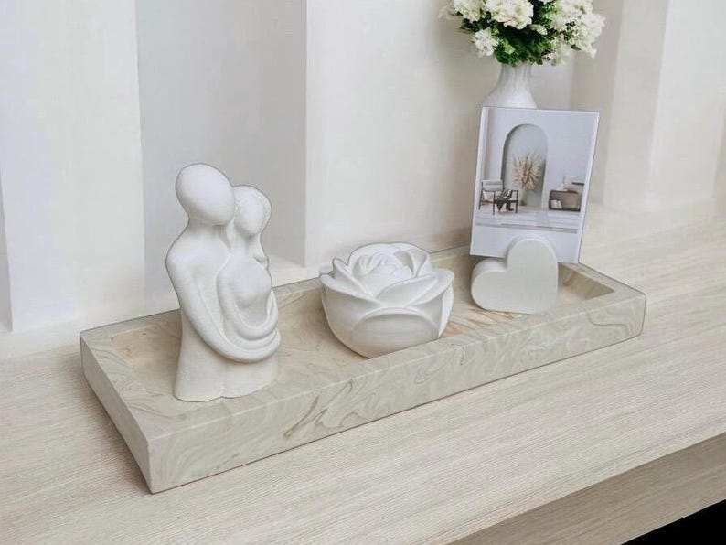 Op de afbeelding: Witte keramische schaal met een wit keramisch beeld van twee figuren die elkaar omhelzen, een witte keramische roos en een wit keramisch hart. De schaal staat op een licht houten oppervlak.
