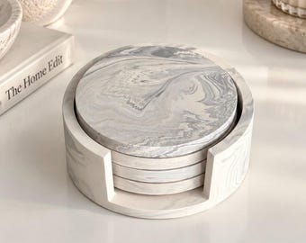 May include: A set of four circular coasters featuring a grey and white marbled pattern, stacked neatly inside a matching cylindrical holder with a front cutout. The coasters and the holder display a consistent swirling stone-like aesthetic.