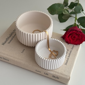 以下が含まれることがあります： 大小2つのオフホワイトの溝付きセラミックボウルが重なっており、金色のジュエリーが入っています。「Love, Life, Style」の文字が入った本と赤いバラも写っています。ボウルは、ホームデコレーションコレクションの一部です。