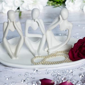 May include: Three white ceramic sculptures of stylized figures sitting on a white oval dish with a pearl necklace and a single red rose.
