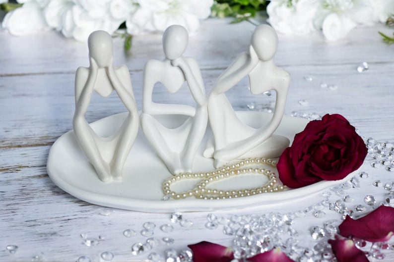 May include: Three white ceramic sculptures of stylized figures sitting on a white oval dish with a pearl necklace and a single red rose.