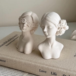 Puede incluir: Dos bustos de mujeres de yeso blanco con detalles florales. Un busto tiene una diadema floral que cubre los ojos, el otro tiene un detalle floral en el lado de la cabeza. Los bustes est&aacute;n sentados sobre un libro con el texto "Home Decor" y "The Art of Neutral Aesthetic Decor" visible.