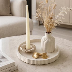 Op de afbeelding: Een rond, gebroken wit dienblad met ribbelpatroon, met daarop een bijpassende geribbelde vaas gevuld met gedroogde beige bloemen, een geribbelde kaarsenhouder met een lange crèmekleurige kaars en een klein, goudkleurig decoratief strikje.