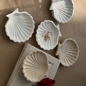 May include: Four white ceramic seashell-shaped trinket dishes on a beige surface. One dish has gold rings inside. A red rose and a book with the text "Style" and "The Neutral Aesthetic" are in the background.