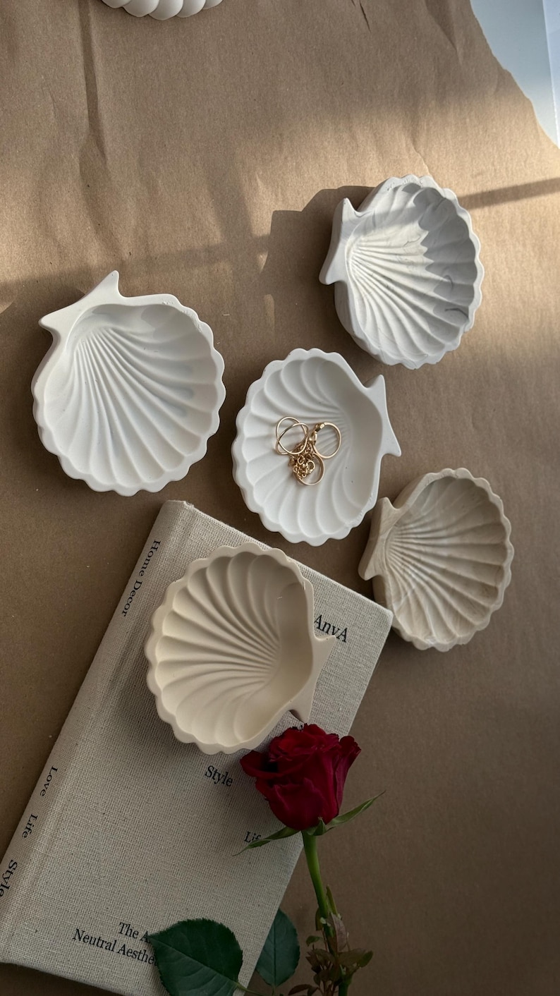May include: Four white ceramic seashell-shaped trinket dishes on a beige surface. One dish has gold rings inside. A red rose and a book with the text "Style" and "The Neutral Aesthetic" are in the background.