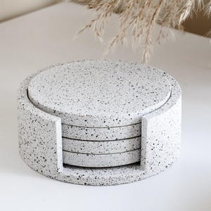 Puede incluir: Un juego de cuatro posavasos redondos y un soporte a juego. Los posavasos y el soporte son de color gris claro con un patr&oacute;n moteado negro. El soporte tiene lados curvos con recortes.