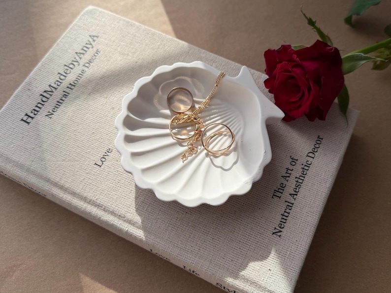 May include: A white ceramic seashell-shaped trinket dish with gold rings and a chain necklace inside. The dish is sitting on a book with the text "HandmadebyAnya Neutral Home Decor" and "The Art of Neutral Aesthetic Decor".