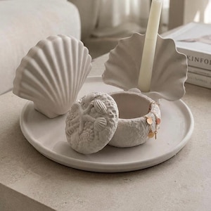 Può includere: Una collezione di oggetti decorativi in ceramica bianca, tra cui un portacandele a forma di conchiglia con una candela bianca, una scultura a conchiglia e un piccolo portagioie rotondo con coperchio. Gli oggetti sono disposti su un vassoio bianco.