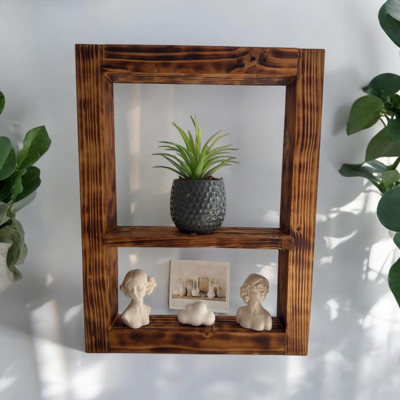 Puede incluir: Una estanter&iacute;a de madera r&uacute;stica con dos estantes, que presenta una peque&ntilde;a planta en maceta, dos figuritas decorativas y una foto enmarcada. La estanter&iacute;a tiene un acabado de madera marr&oacute;n oscuro y est&aacute; hecha de madera reciclada.