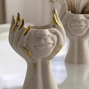 Pode incluir: Um par de vasos de cerâmica branco-sujo com um design de rosto. Os vasos apresentam mãos segurando o rosto, com detalhes em folha de ouro nas pontas dos dedos e no rosto. Um vaso está em foco, o outro está desfocado no fundo.