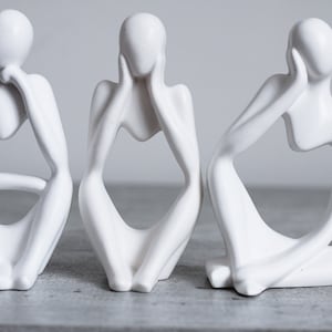 May include: Three white ceramic sculptures of abstract figures in a seated pose. Each figure has its hands covering its face, eyes, or mouth.