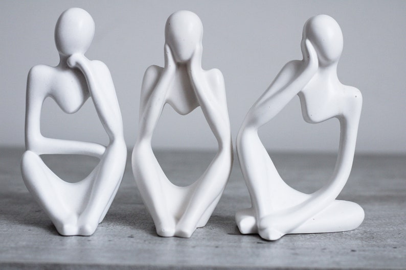 May include: Three white ceramic sculptures of abstract figures in a seated pose. Each figure has its hands covering its face, eyes, or mouth.