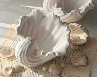 May include: Two large, white ceramic seashell-shaped bowls with ruffled edges. Several real seashells and pearls are scattered around the bowls on a piece of aged paper. The bowls have a glossy finish and are arranged on a light-colored surface.