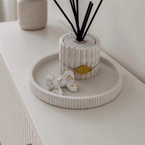 Puede incluir: Un difusor de cerámica blanca acanalada con varillas negras, colocado sobre una bandeja a juego. Un lazo decorativo con detalles de pan de oro está en la bandeja. Un pequeño jarrón beige con una planta está en el fondo.