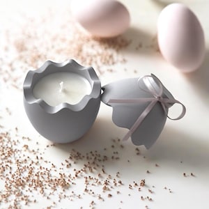 May include: A grey egg-shaped candle holder with a white candle inside. The holder is open and the top half is lying next to it. The top half has a grey ribbon tied around it.