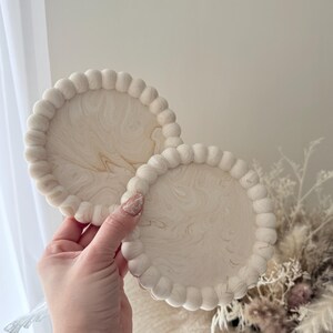 May include: Two round, off-white coasters with a beaded edge and a marbled, cream-colored design. The coasters are held in a hand, showcasing their decorative and functional nature. They are suitable for home decor.