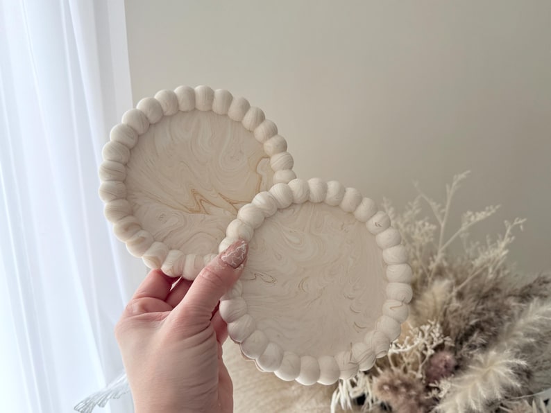 May include: Two round, off-white coasters with a beaded edge and a marbled, cream-colored design. The coasters are held in a hand, showcasing their decorative and functional nature. They are suitable for home decor.
