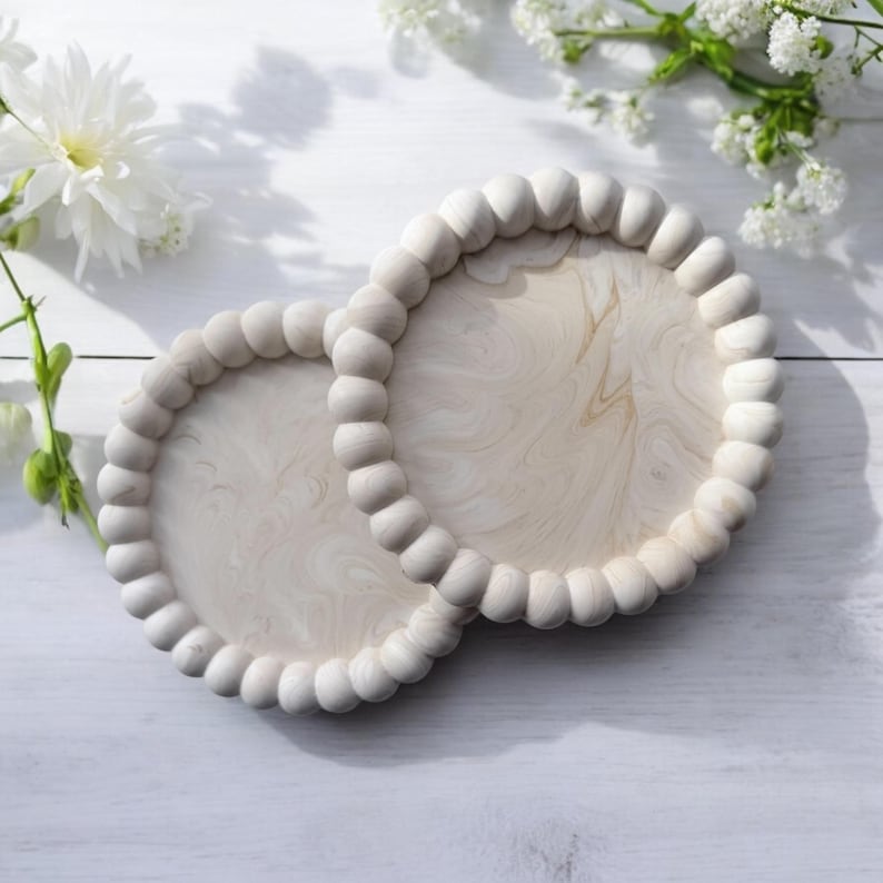May include: Two round, light-colored wooden coasters with a beaded edge. The coasters have a light wood grain pattern and are set on a white surface, with white flowers in the background.