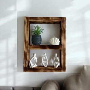 May include: A rustic wooden shelf with two shelves, featuring a small potted plant, a white seashell, and three white ceramic figurines. The shelf is mounted on a white wall.