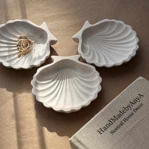 May include: Three white ceramic seashell-shaped dishes. The dishes are arranged on a light brown surface. The dish in the centre is slightly larger than the other two. The dishes are perfect for holding jewellery or other small items.