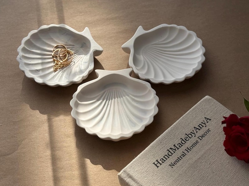 May include: Three white ceramic seashell-shaped dishes. The dishes are arranged on a light brown surface. The dish in the centre is slightly larger than the other two. The dishes are perfect for holding jewellery or other small items.