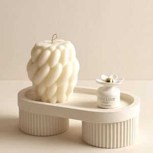 Puede incluir: Una vela trenzada de color crema con una mecha dorada se encuentra sobre una bandeja blanca de forma ovalada con un pequeño difusor blanco en forma de flor. El difusor tiene el texto "TIRS LOVE". La bandeja tiene un diseño acanalado.