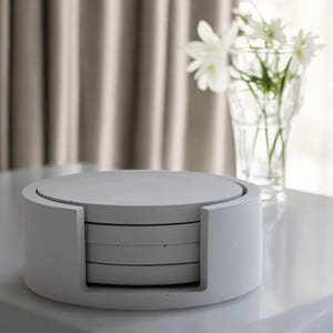 Puede incluir: Un juego de cuatro posavasos redondos de color gris claro en un soporte a juego. Los posavasos est&aacute;n apilados ordenadamente, con un acabado liso y mate. El soporte tiene un dise&ntilde;o curvo, que facilita el acceso a los posavasos. El juego est&aacute; sobre una superficie blanca.