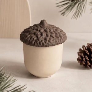 May include: A ceramic container shaped like an acorn, featuring a smooth, light beige base and a textured, dark brown lid with a sculpted cap. The object has a rounded, cylindrical body and a detailed, bumpy surface on the top portion.
