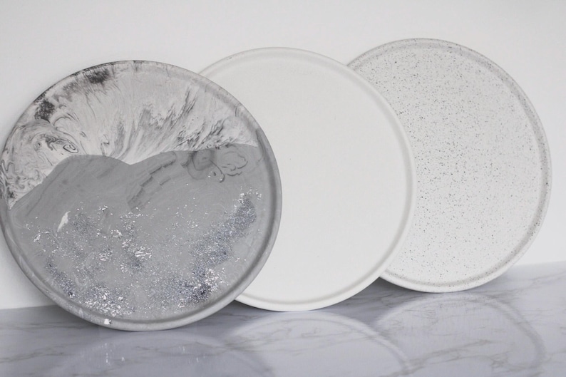May include: Three round concrete trays with different finishes. One tray has a white speckled finish, one has a white finish, and one has a gray and white marbled finish.