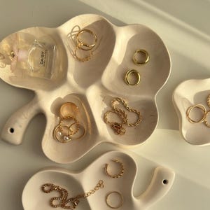 May include: A collection of gold jewelry, including rings, earrings, and a necklace, displayed on off-white, leaf-shaped and free-form ceramic trays. A small perfume bottle is also present. The jewelry is arranged to showcase its design and style.