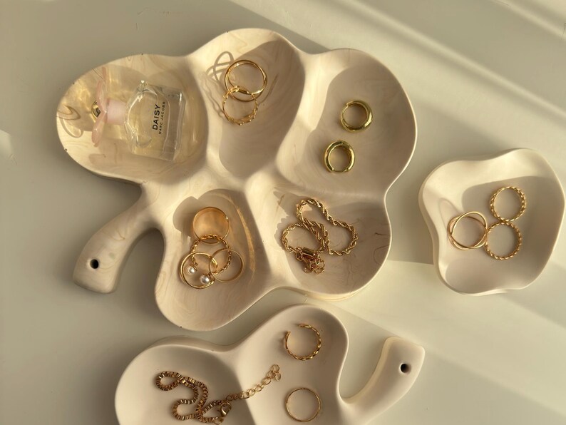 May include: A collection of gold jewelry, including rings, earrings, and a necklace, displayed on off-white, leaf-shaped and free-form ceramic trays. A small perfume bottle is also present. The jewelry is arranged to showcase its design and style.