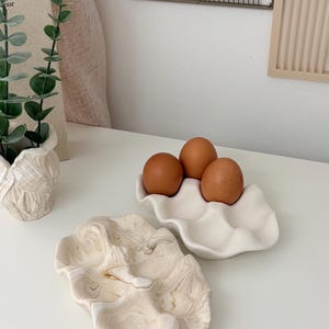 May include: A still-life composition featuring three brown eggs in a white, wavy ceramic egg holder. A second, decorative, clam-shaped dish with a marbled beige and white pattern is also present. The scene is set on a white surface.