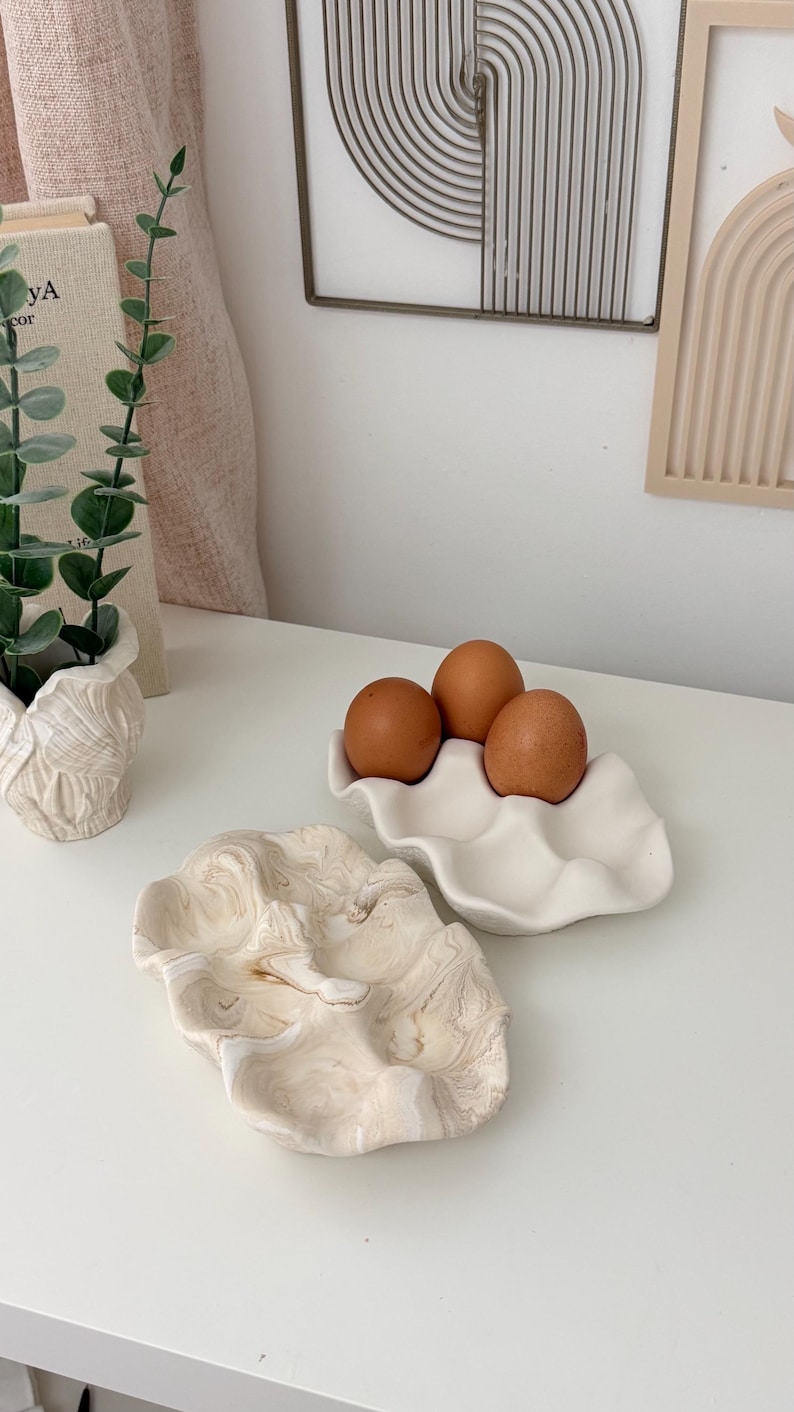 May include: A still-life composition featuring three brown eggs in a white, wavy ceramic egg holder. A second, decorative, clam-shaped dish with a marbled beige and white pattern is also present. The scene is set on a white surface.