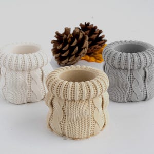 May include: Three ceramic planters shaped like knitted sweaters in white, grey, and beige. The planters are sitting on a white surface.