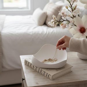 May include: A white jewellery dish, holding a gold necklace, sits on a book with the word "NEESTIETIC" on the cover. The scene is set on a wooden bedside table, with a bed and floral arrangement in the background.