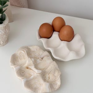 May include: Two ceramic egg holders. One is white and holds three brown eggs. The other is a marbled beige and white design. Both are on a white surface. The egg holders are decorative and functional.
