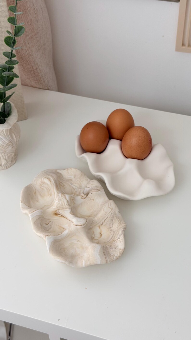 May include: Two ceramic egg holders. One is white and holds three brown eggs. The other is a marbled beige and white design. Both are on a white surface. The egg holders are decorative and functional.