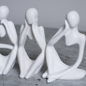 May include: Three white ceramic sculptures of abstract figures in a seated pose, each with their hand on their face.