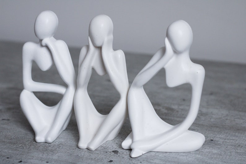 May include: Three white ceramic sculptures of abstract figures in a seated pose, each with their hand on their face.