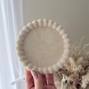May include: A round, off-white decorative tray with a beaded edge. The tray's surface has a marbled pattern with subtle gold veining. The tray is held in a hand, with a soft, neutral background.