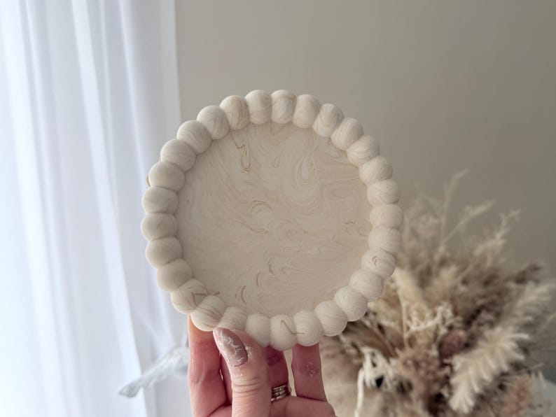 May include: A round, off-white decorative tray with a beaded edge. The tray's surface has a marbled pattern with subtle gold veining. The tray is held in a hand, with a soft, neutral background.