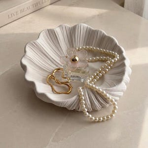 Peut inclure: Un plateau décoratif blanc en forme de coquillage contient un collier de perles, une bague dorée et un flacon de parfum avec un bouchon en forme de fleur. Le plateau est posé sur une surface claire, avec un livre qui dit "LIVE BEAUTIFULLY" en arrière-plan.