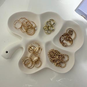 May include: A white, clover-shaped jewelry dish filled with various gold rings and a gold chain. The rings are in different styles, including bands, twisted designs, and rings with small embellishments. A small, iridescent box is in the background.