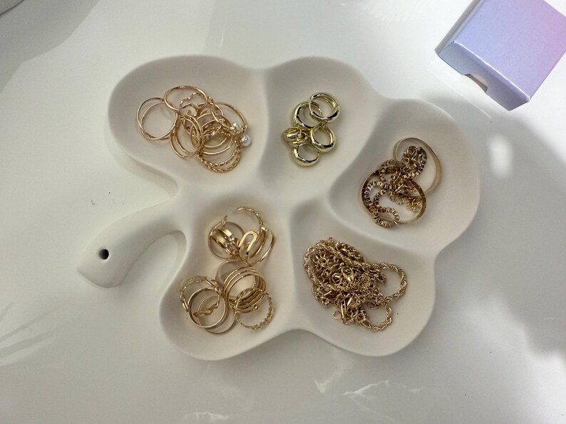 May include: A white, clover-shaped jewelry dish filled with various gold rings and a gold chain. The rings are in different styles, including bands, twisted designs, and rings with small embellishments. A small, iridescent box is in the background.