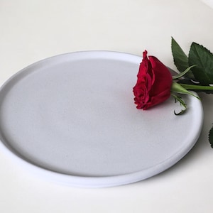May include: A white ceramic plate with a single red rose on top. The plate is round and has a slightly raised edge.