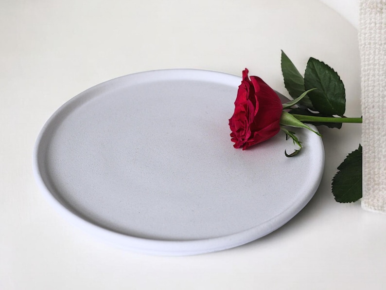 May include: A white ceramic plate with a single red rose on top. The plate is round and has a slightly raised edge.