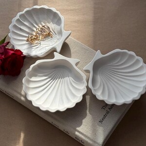 May include: Three white ceramic seashell-shaped trinket dishes. The dishes are arranged on a book with the text "The Neutral Aesthetic" visible.