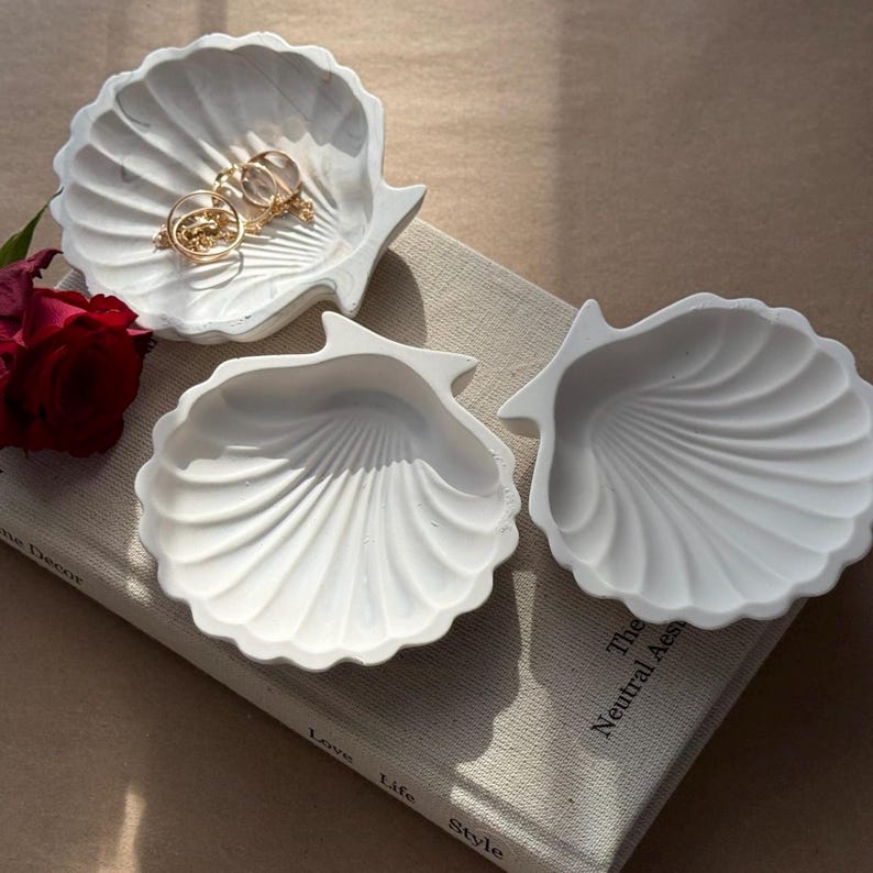 May include: Three white ceramic seashell-shaped trinket dishes. The dishes are arranged on a book with the text "The Neutral Aesthetic" visible.