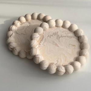 Pode incluir: Duas bandejas ovais de madeira com um design marmorizado bege claro. Cada bandeja tem uma borda de esferas de madeira. As bandejas são feitas de madeira clara e estão sobre um fundo branco.